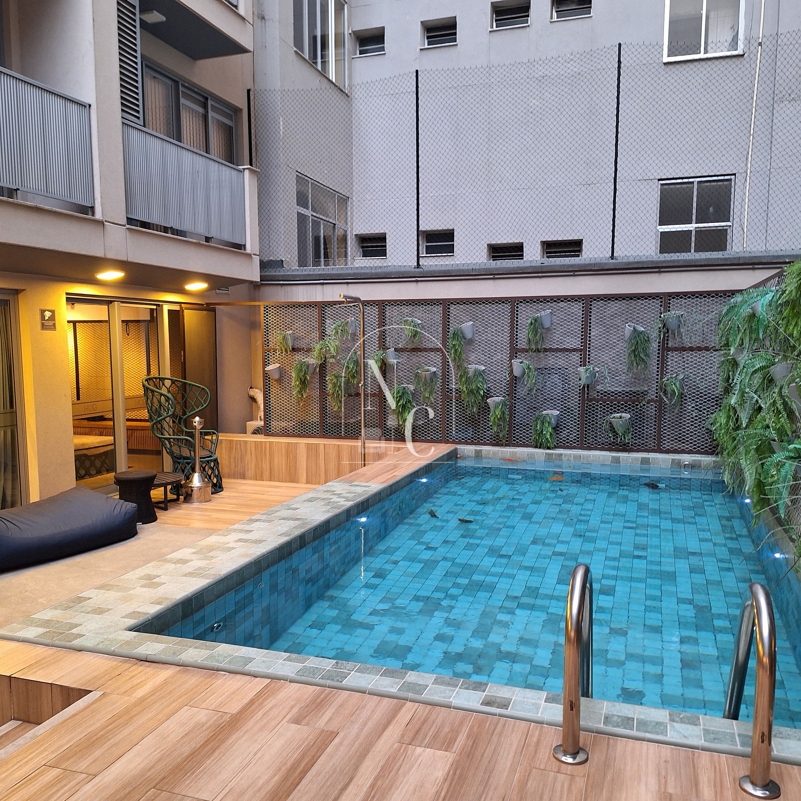 A swimming pool with a wooden deck and metal railings, surrounded by a building with balconies and a mesh fence.
