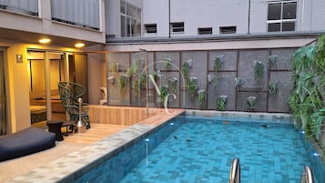Ein Schwimmbad mit Holzterrasse und Metallgeländern, umgeben von einem Gebäude mit Balkonen und einem Maschendrahtzaun.