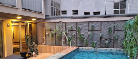 A swimming pool with a wooden deck and metal railings, surrounded by a building with balconies and a mesh fence.