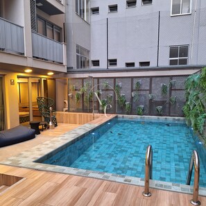 Una piscina con cubierta de madera y barandillas metálicas, rodeada por un edificio con balcones y una valla de malla.