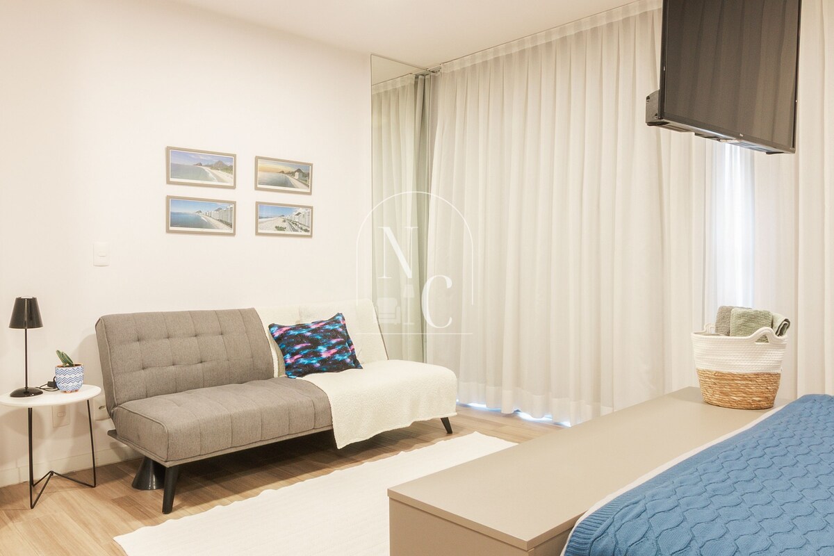 Un dormitorio moderno con sofá, cama, televisor y una mesita de noche con lámpara y planta.