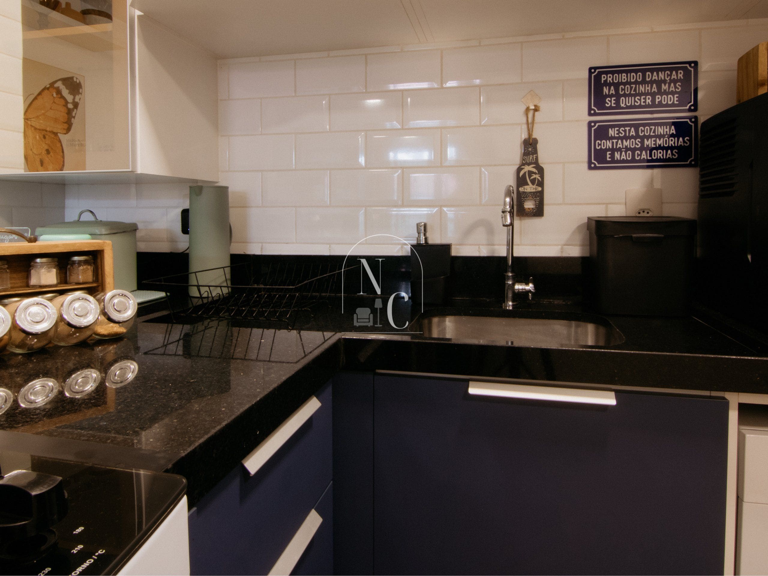 A kitchen with white tiled backsplash, a black countertop, a stainless steel sink, and a coffee machine. There are jars on the counter and a sign on the wall.