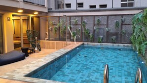 Una piscina amb un solàrium de fusta i baranes metàl·liques, envoltada per un edifici amb balcons i una tanca de malla.