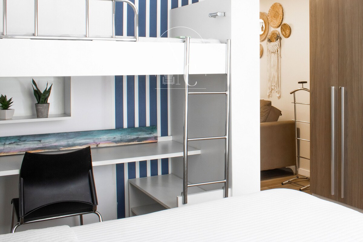 Una cama individual superpuesta con escritorio, una silla, una planta pequeña y una pared con rayas verticales.