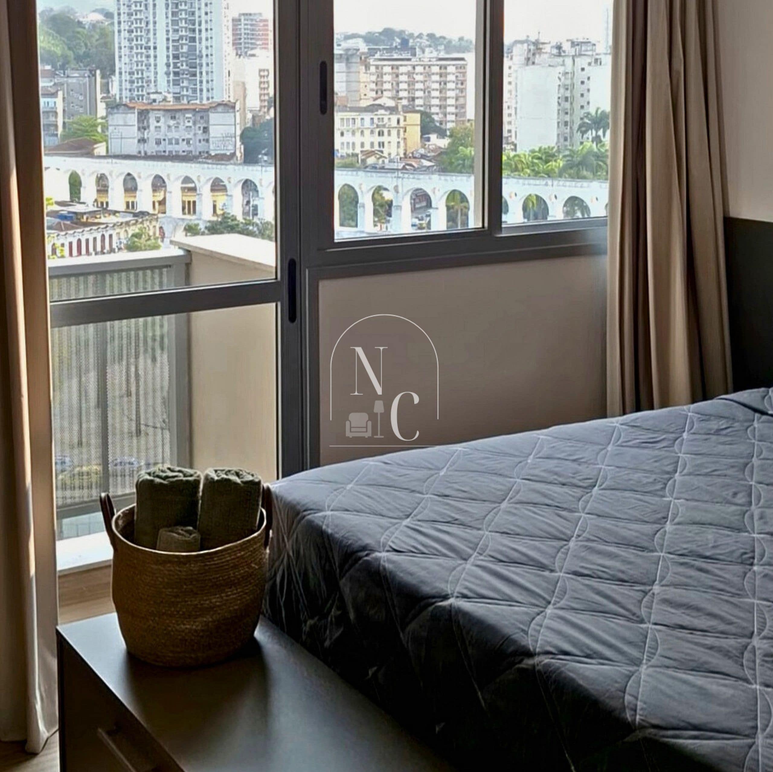 A bedroom with a view of a cityscape through a window, a bed with a quilt, a basket on a nightstand, and a logo on the wall.
