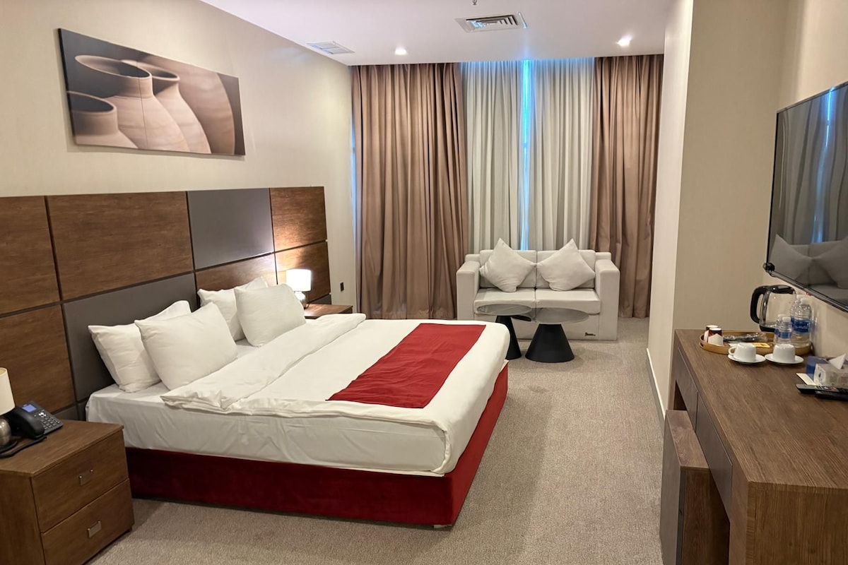 A hotel room with a large bed, a sofa, a TV, and a desk with a tray of items.