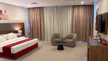 Una habitación de hotel con cama, dos sillones, una mesa de centro, televisor y un amplio ventanal con cortinas.