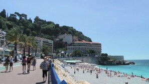 A coastal promenade with a beach, palm trees, and buildings in the background. - Studio à louer Carré d'Or à Nice (Nice)