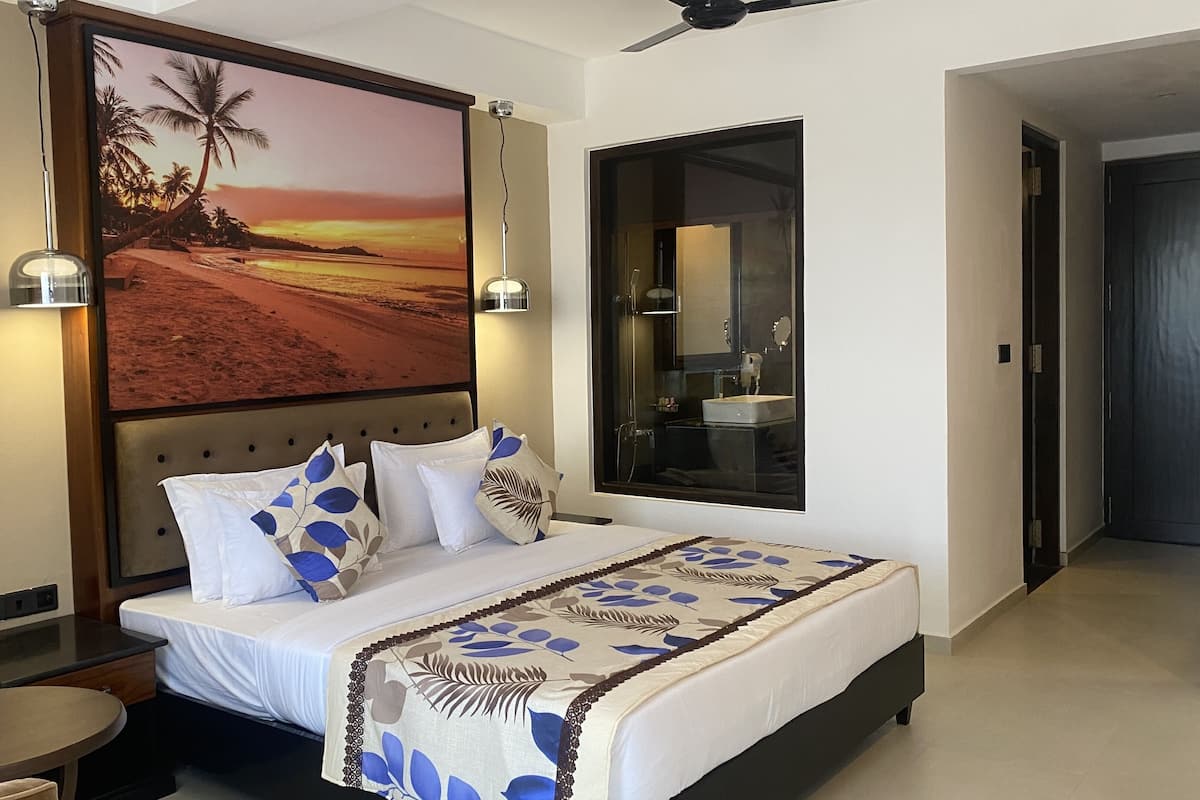 A hotel room with a bed, a nightstand, a desk, a chair, a large painting of a beach scene, a mirror, and a door.