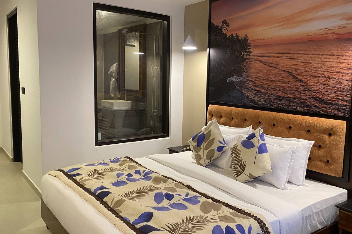 A bedroom with a large bed, a headboard, a window, and a wall-mounted artwork of a beach scene.