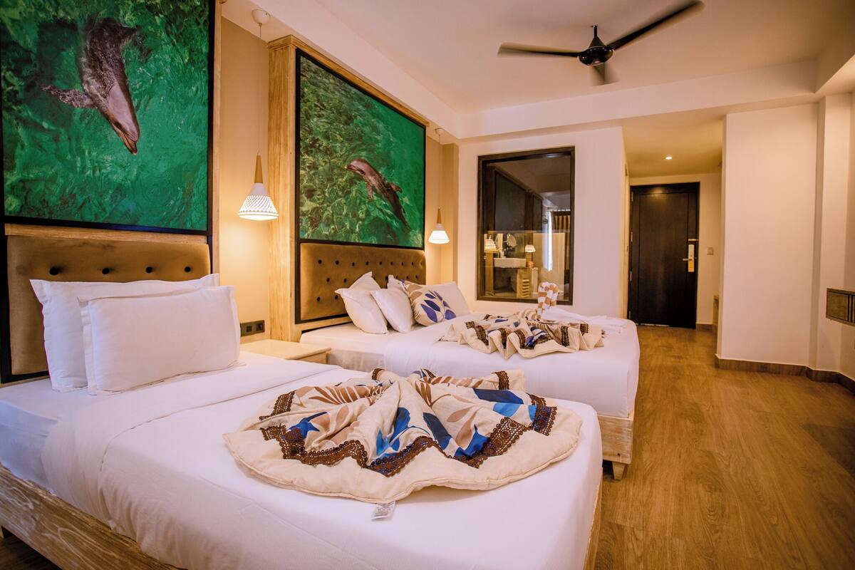 A hotel room with two beds, a ceiling fan, artwork on the walls, and a view into the bathroom.