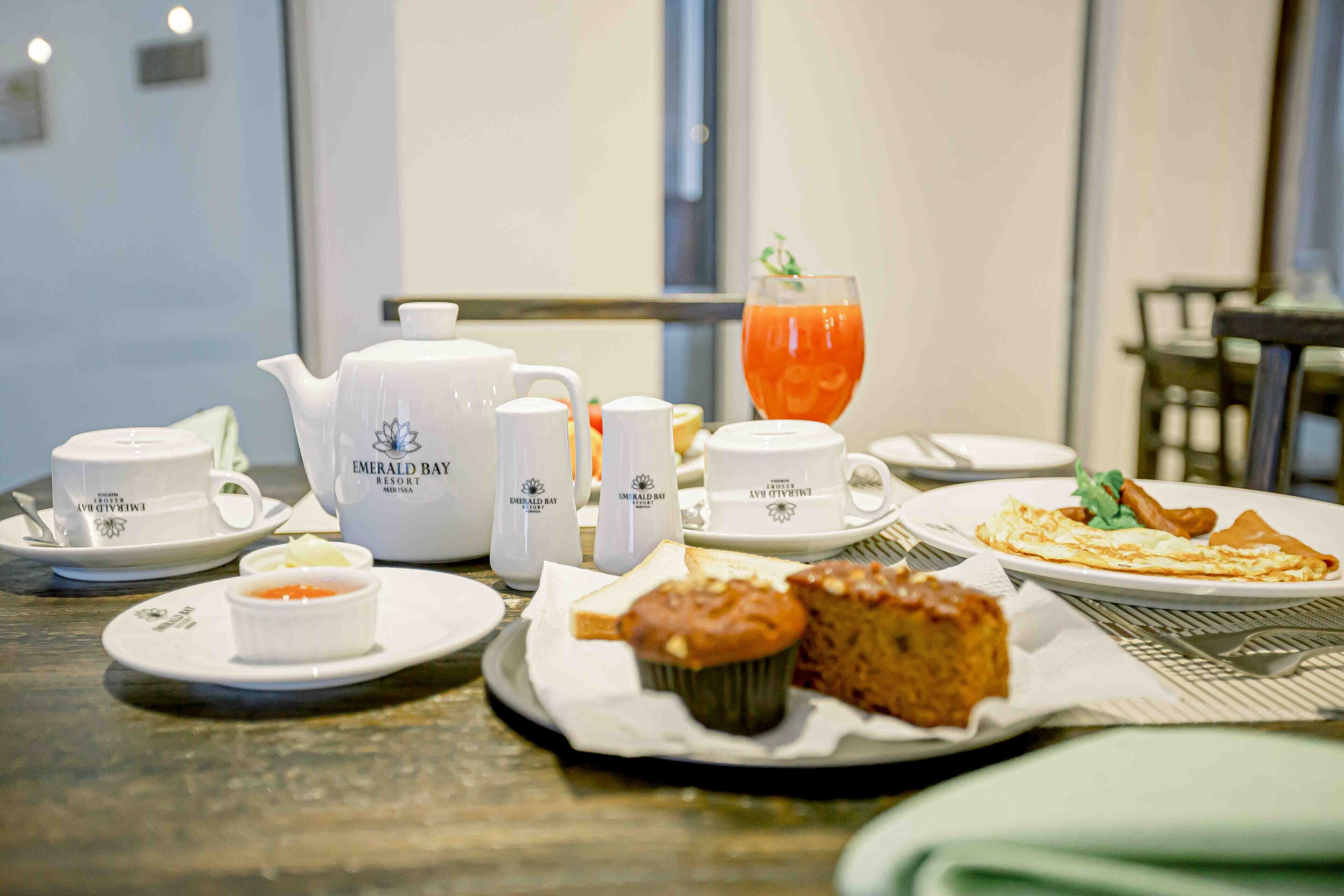A breakfast setting with tea service, a glass of orange juice, muffins, and a plate of eggs and sausage.