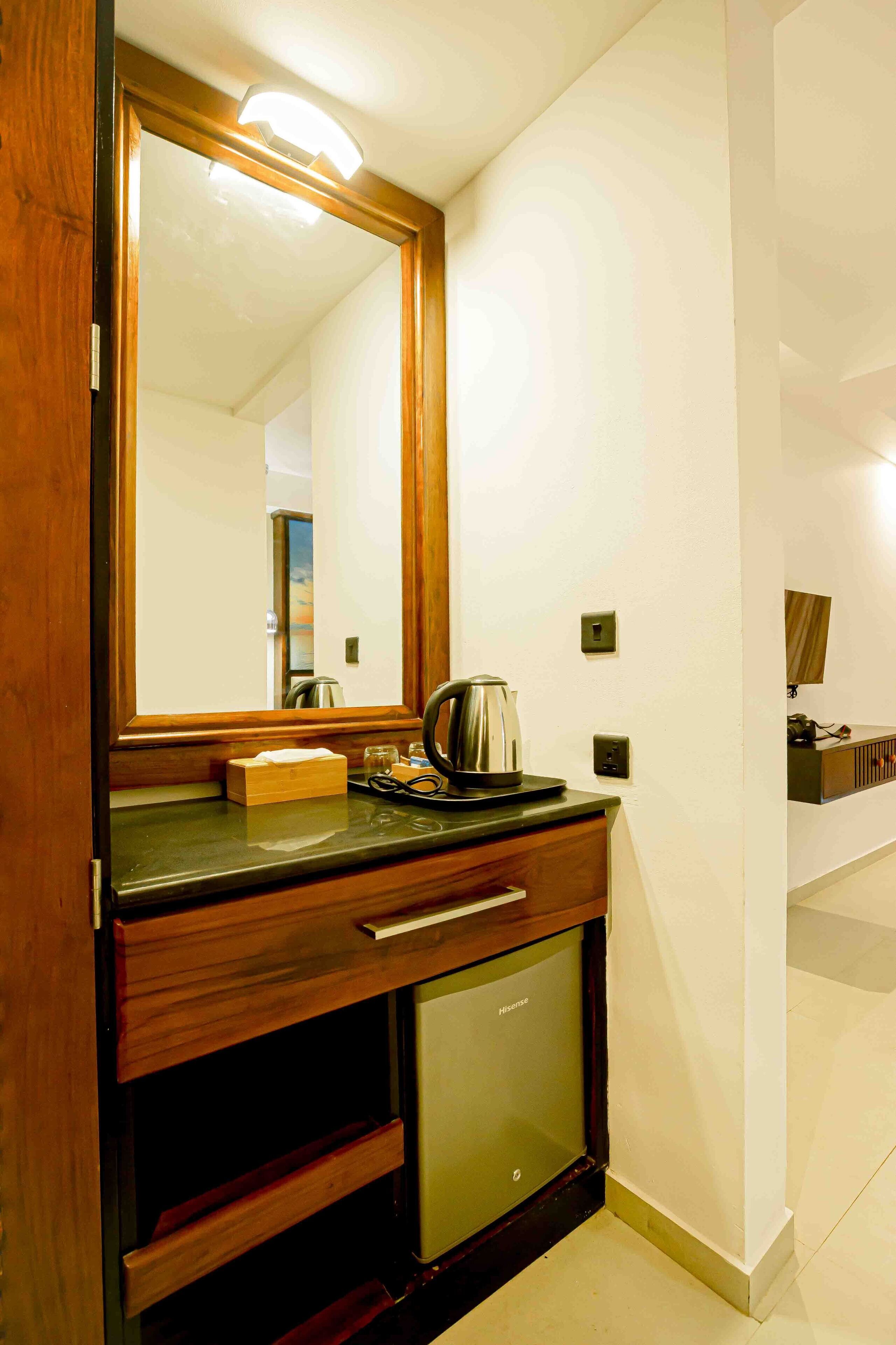 A hotel room with a wooden vanity, a mirror, a small refrigerator, and a countertop with a kettle and a box.