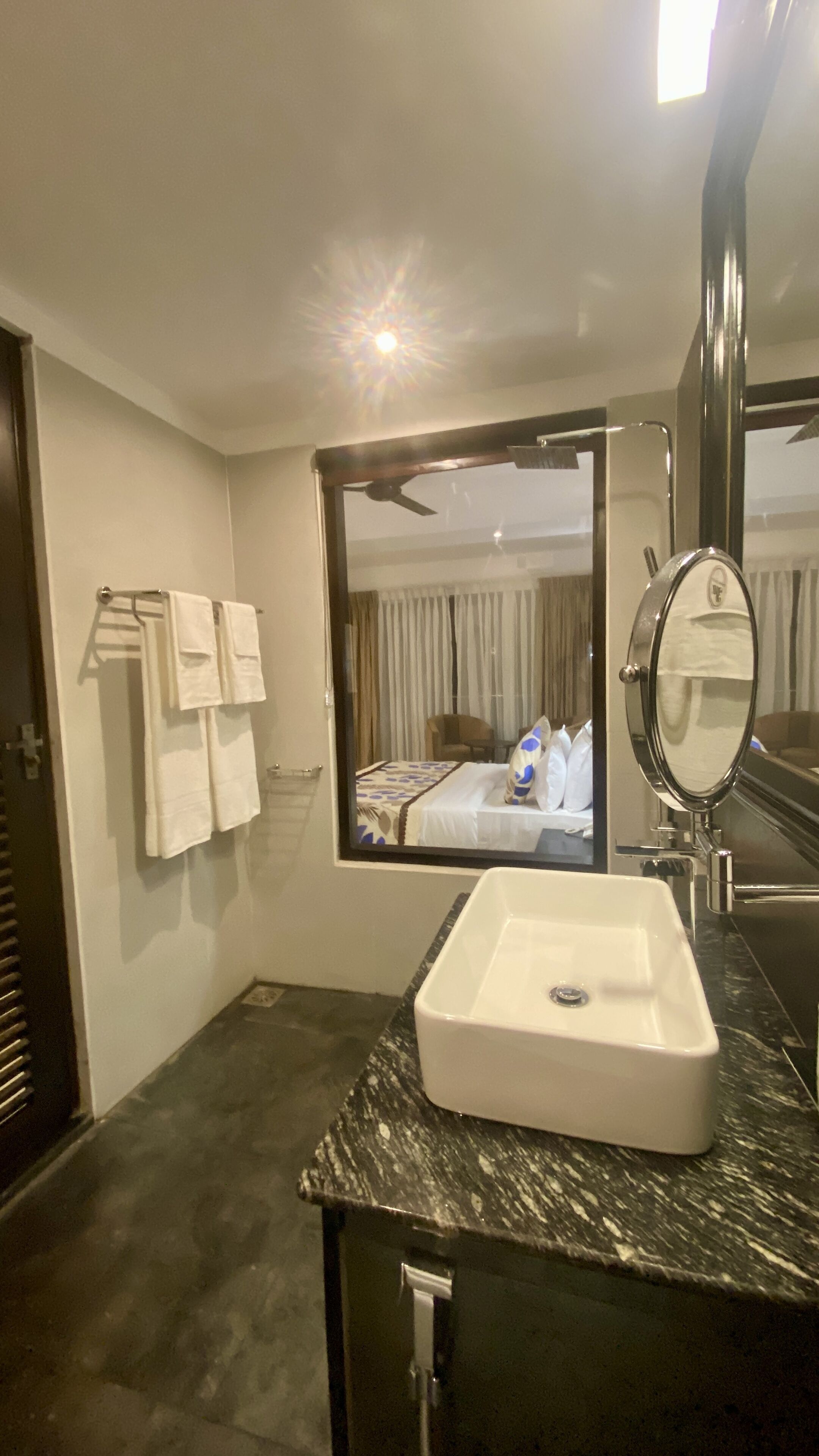 Un baño moderno con encimera de mármol, un lavabo blanco, un espejo grande y vistas a una cama con ventilador de techo.