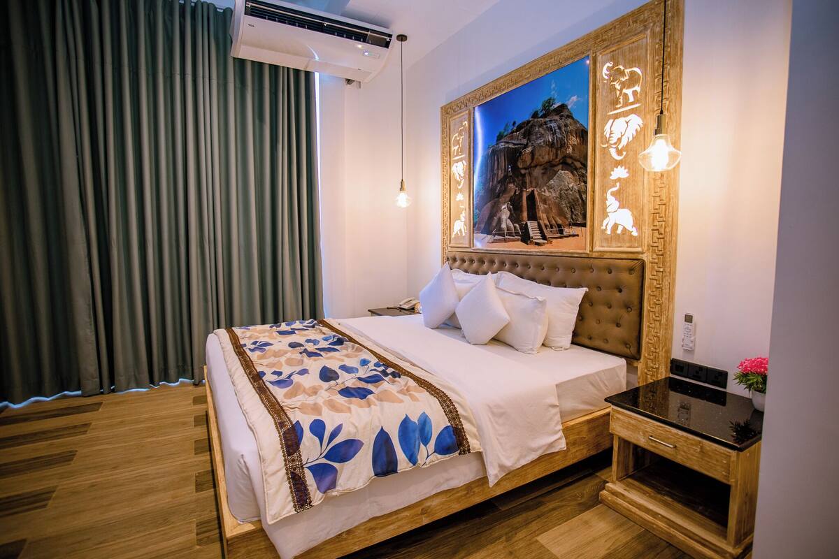 A bedroom with a bed, a nightstand, an air conditioner, a wall-mounted light fixture, and a framed picture above the bed.