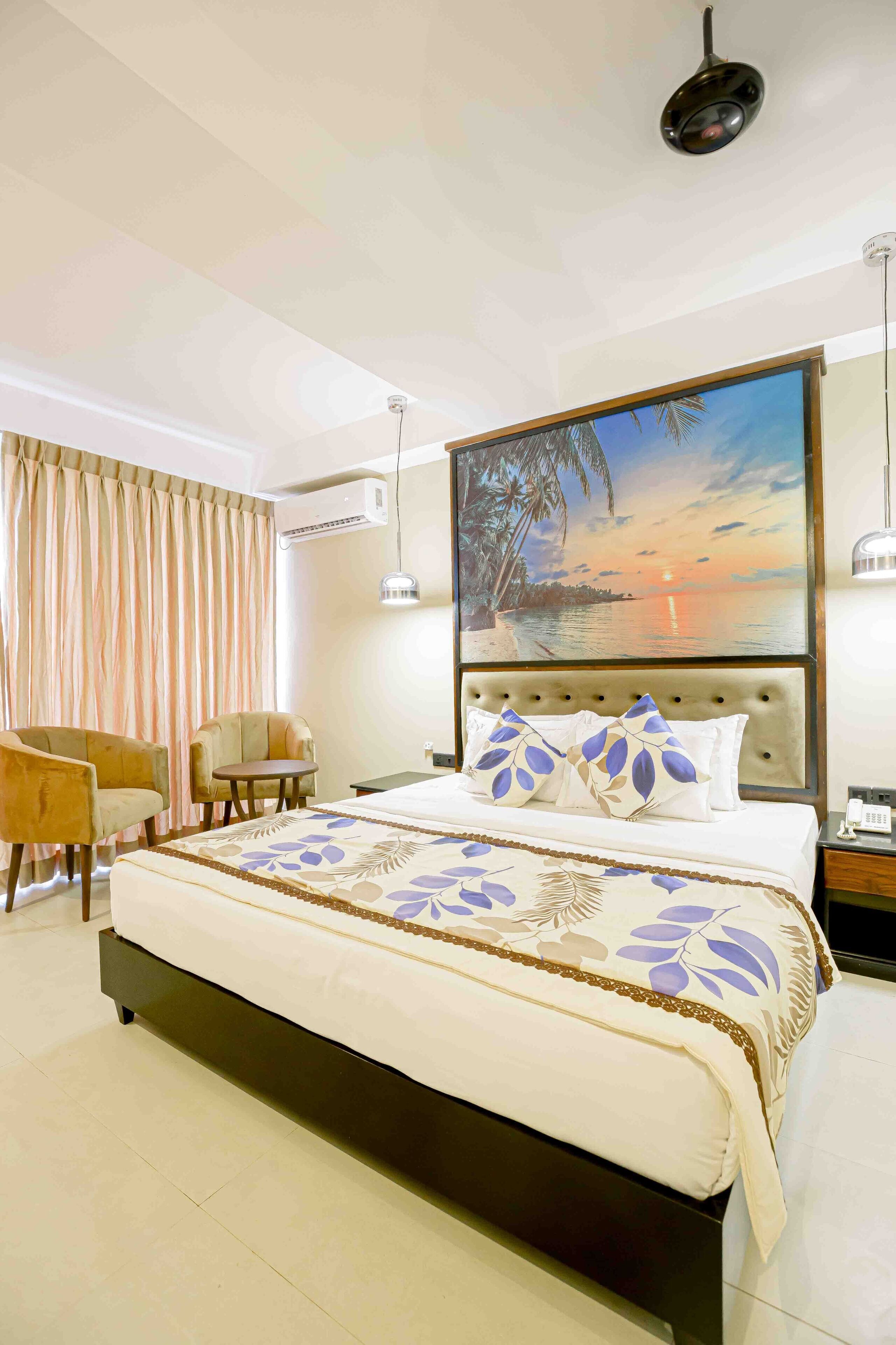 Une chambre d’hôtel avec un grand lit, deux fauteuils, une petite table et un tableau représentant un coucher de soleil sur l’eau.
