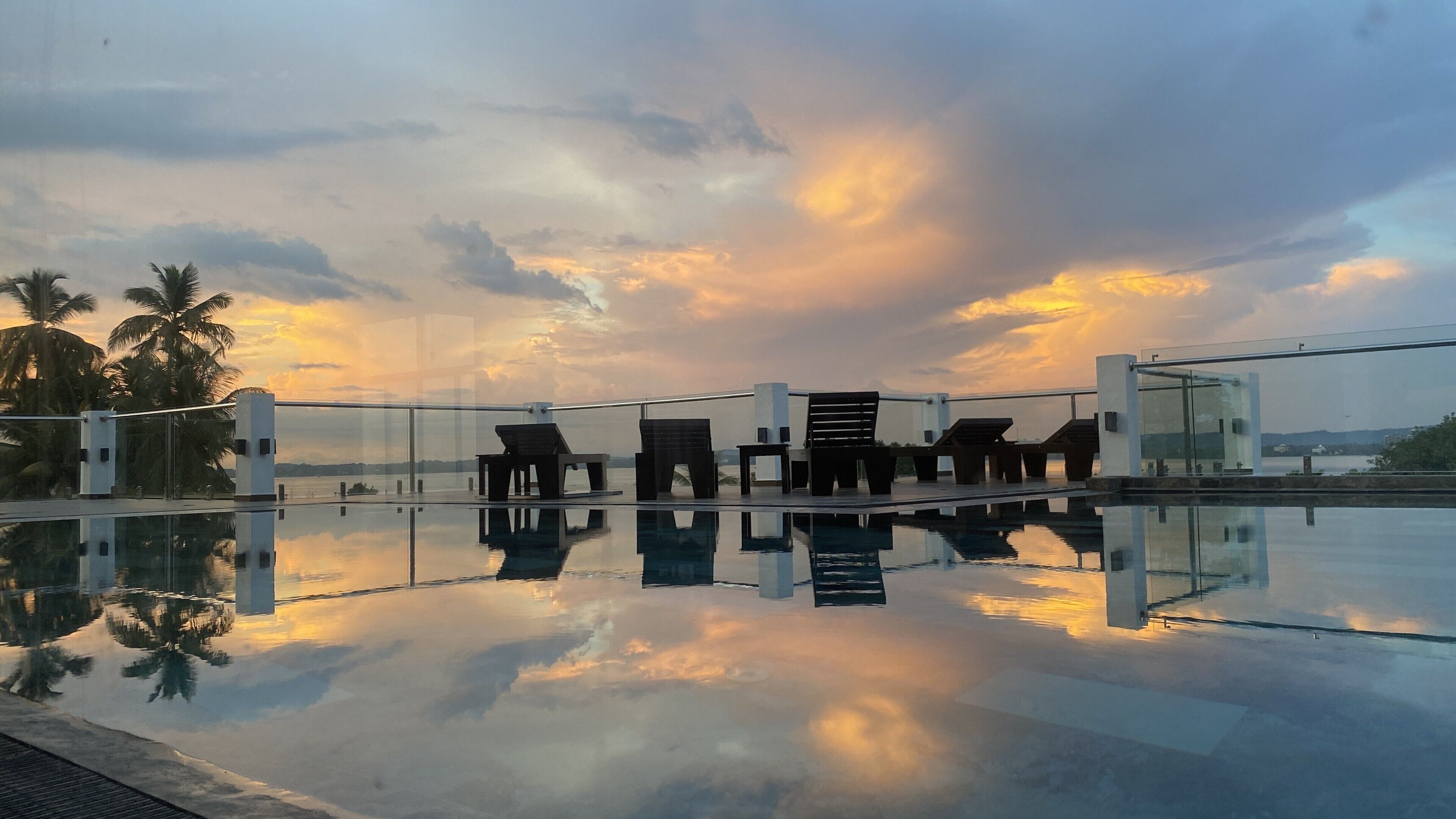 A modern outdoor seating area with a glass balustrade overlooking a body of water, palm trees, and a sunset sky.