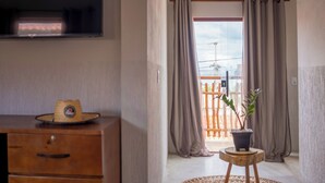Una habitación con un aparador de madera, una planta en maceta sobre un taburete y vistas a un balcón con cortinas.