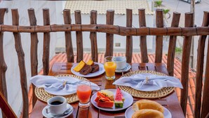 Una mesa de madera con desayuno, incluyendo pan, fruta y bebidas, en un balcón con vistas al mar.