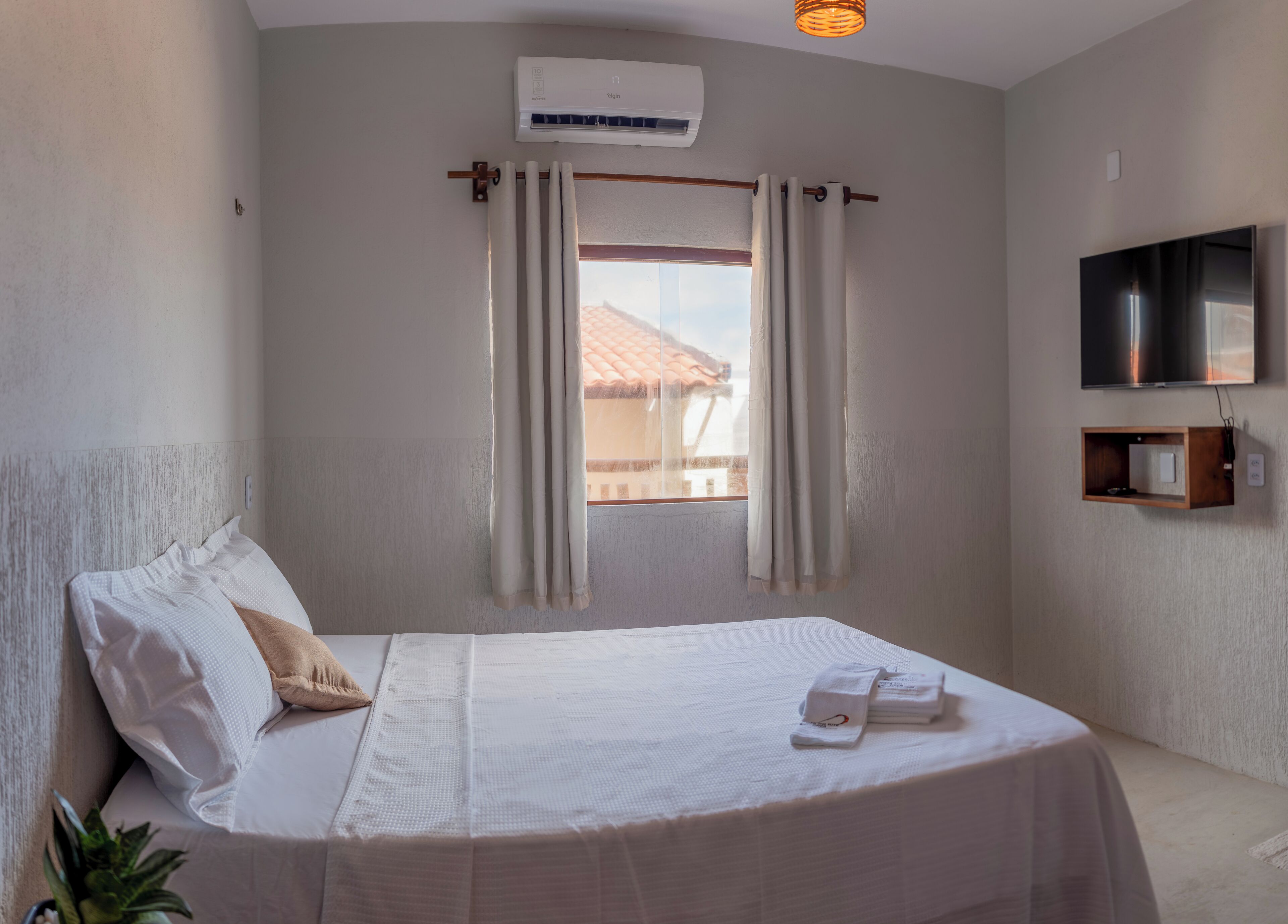 Een netjes opgemaakt bed met witte lakens en een beige kussen, een aan de muur bevestigde televisie, een klein houten plankje en een airconditioner boven een raam met gordijnen.