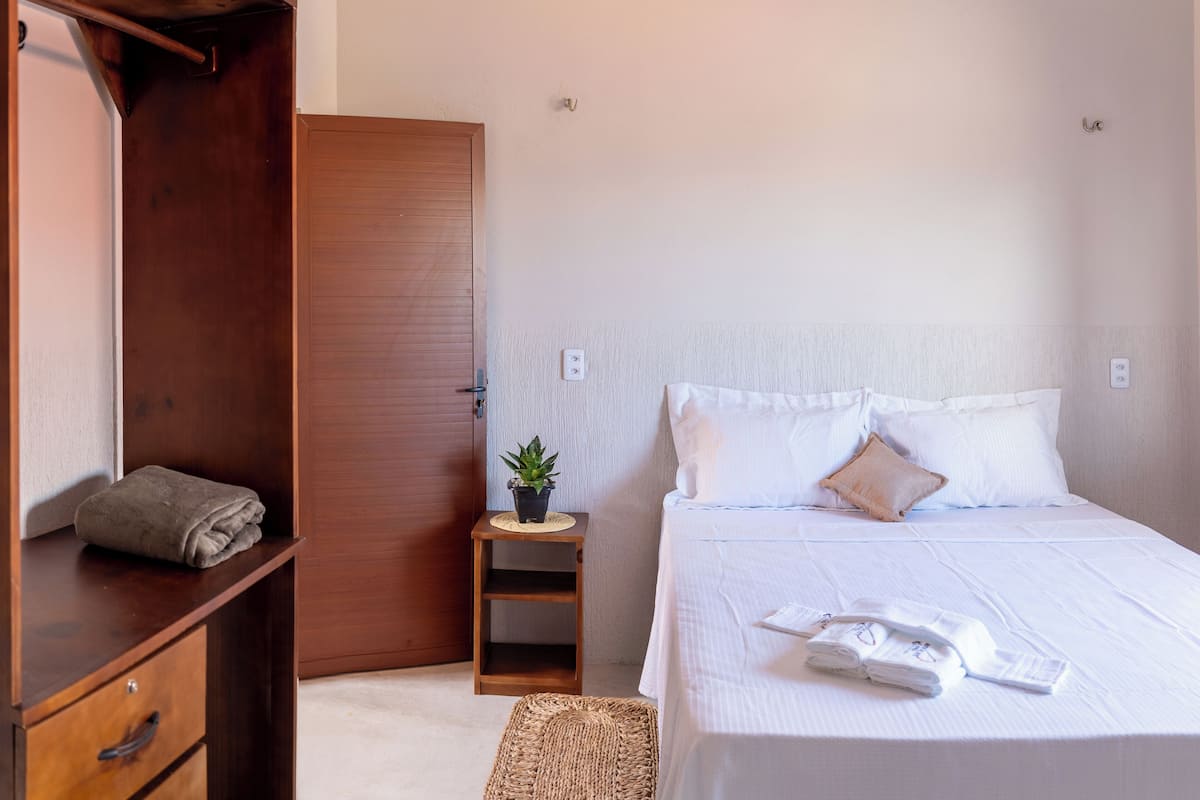 A neatly arranged bedroom with a wooden wardrobe, a small bedside table with a plant, and a neatly made bed with white linens and a pillow.