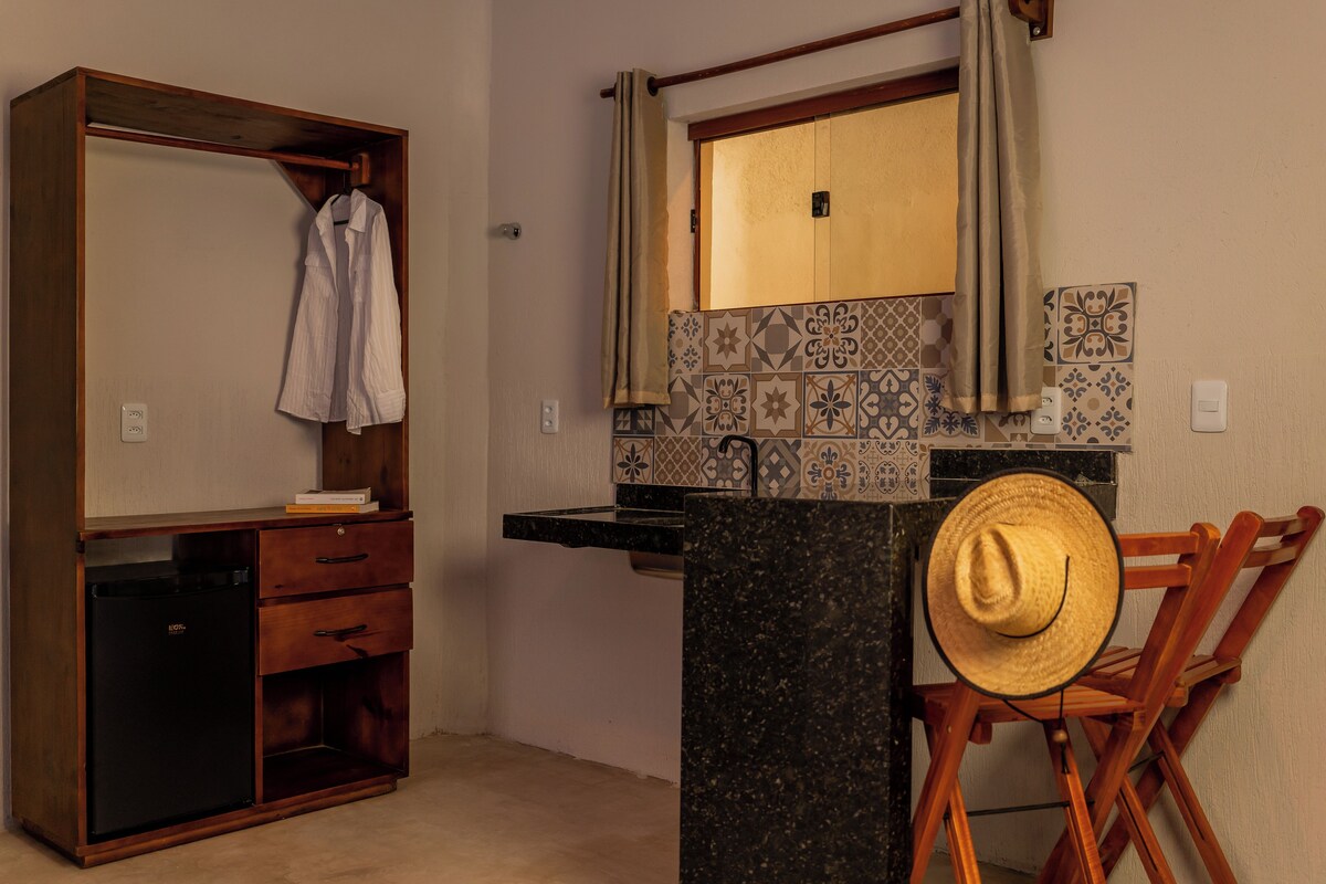 Una habitación compacta con un armario de madera, un escritorio pequeño con cajones, un espejo sobre un salpicadero de azulejos y un sombrero de paja en una silla.
