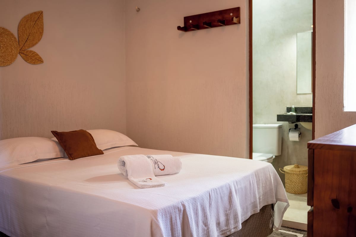 A neatly made bed with a white bedspread, a brown pillow, and a towel. There is a wooden shelf above the bed and a woven wall decoration.