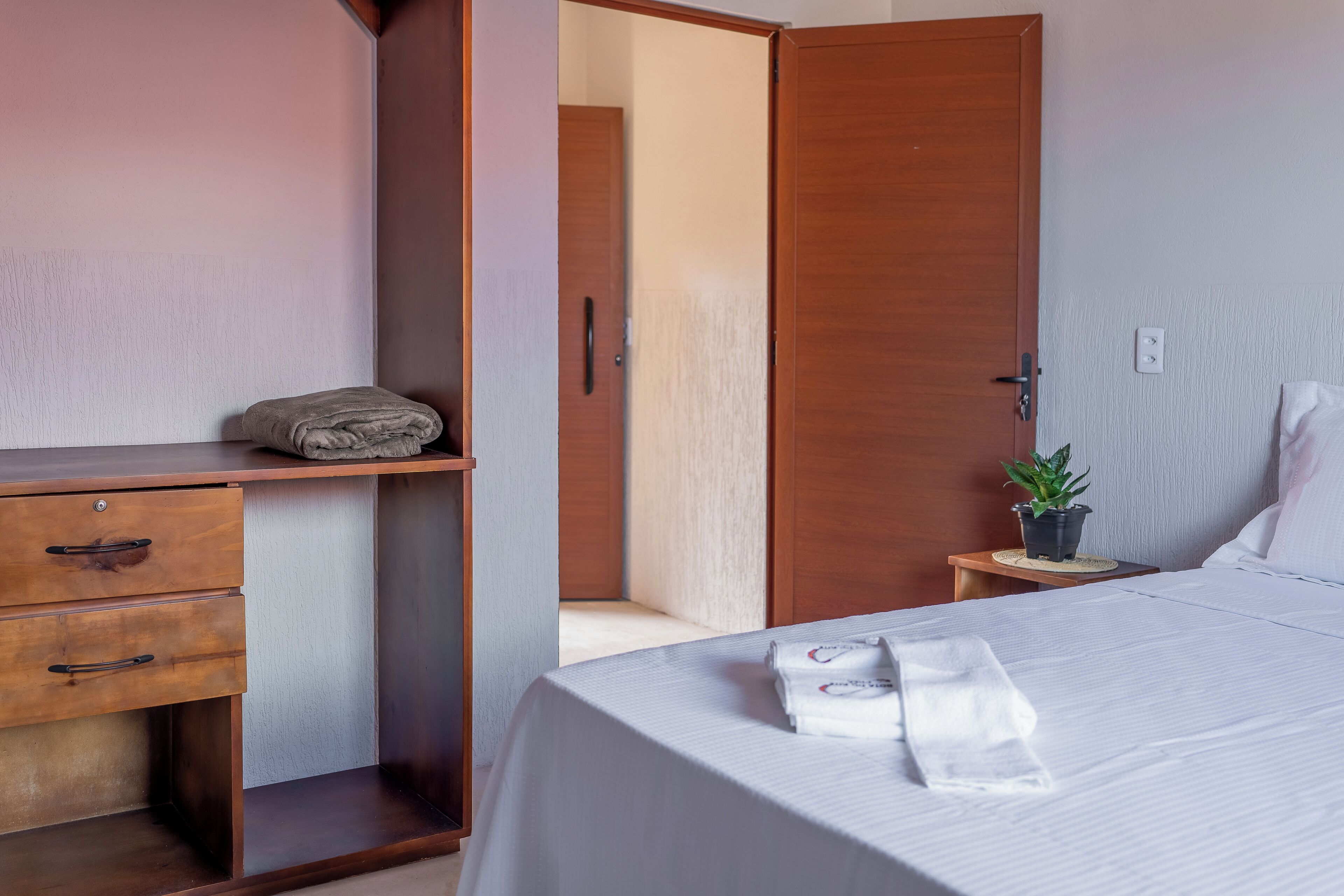 Habitación de hotel con cama, un escritorio de madera con cajones, una mesita con una planta y una puerta que da a otra habitación.