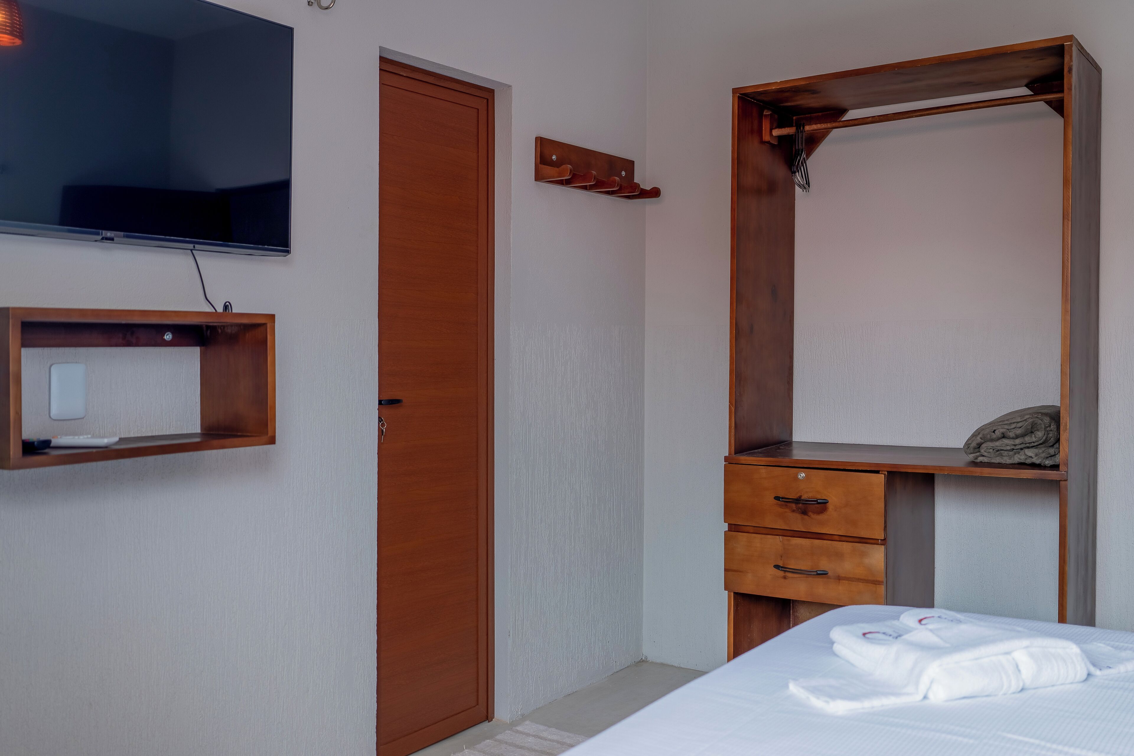 Habitación de hotel con una cama, un armario de madera, un televisor montado en la pared y un pequeño estante con una toalla.