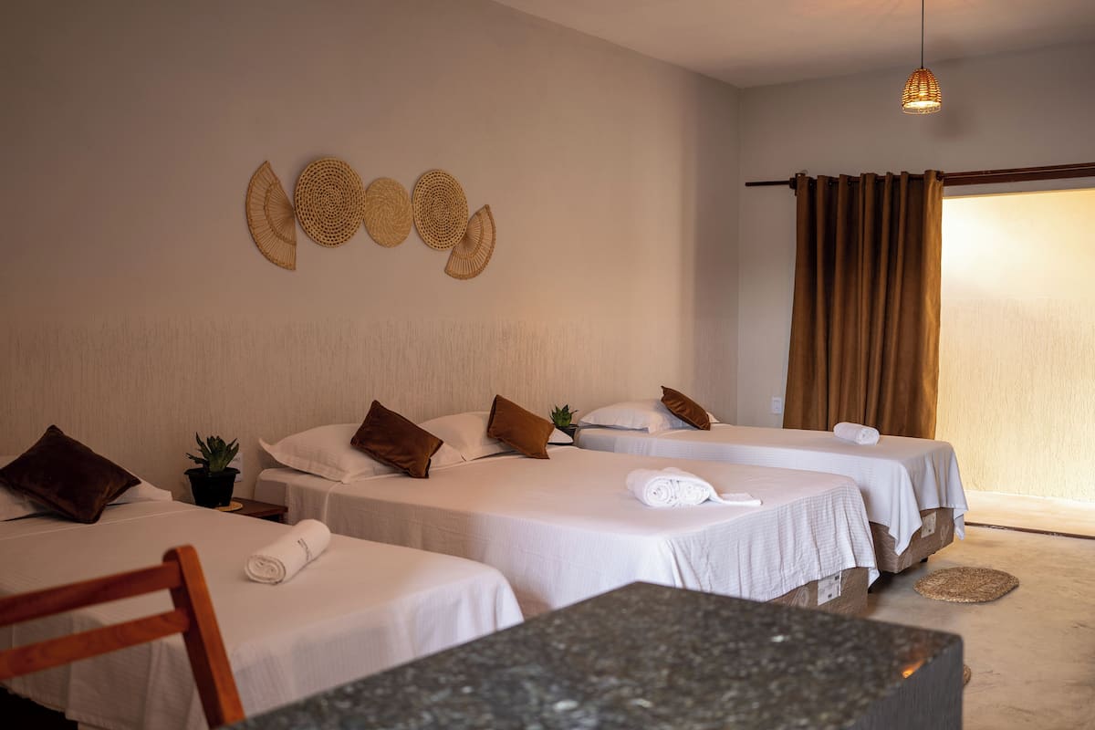 A hotel room with two beds, a small table, a chair, a potted plant, and a wall with woven wall art.