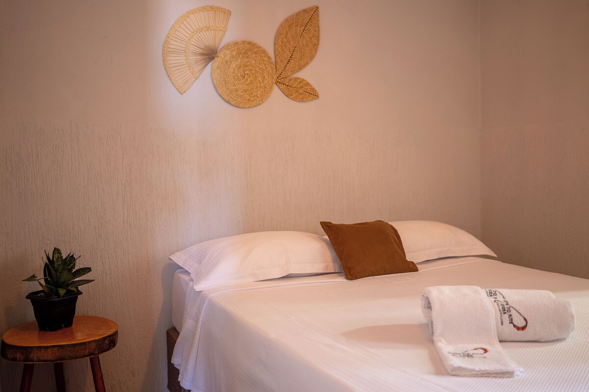 A neatly made bed with white linens, a brown pillow, and a folded towel on the bed. There is a small round wooden stool with a potted plant beside the bed. A decorative wall piece resembling a butterfly is above the bed.