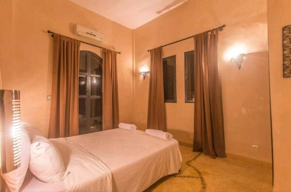 A hotel room with a bed, curtains, an air conditioning unit, and a wall-mounted light fixture.
