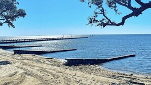 Una playa con un muelle que se adentra en el agua, arena y un gran árbol a la derecha.
