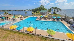 Una piscina con múltiples carriles, rodeada de sombrillas y sillones reclinables, con vista al agua y la costa lejana.