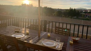 Ein Holztisch für zwei Personen gedeckt mit Tellern, Besteck und Kerzen, unter einem großen Sonnenschirm auf einem Balkon mit Blick auf eine Landschaft aus Bäumen und Gebäuden.