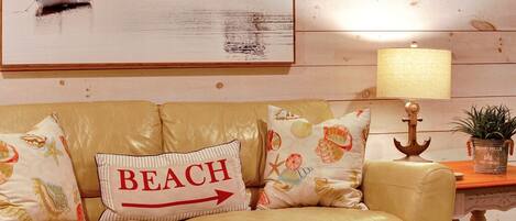Un sofá de cuero color beige con almohadones decorativos, un cojín con motivos playeros, una lámpara y un cuadro enmarcado de una escena de playa.
