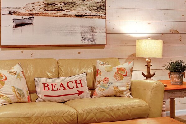 Um sofá de couro bege com almofadas decorativas, uma almofada com tema de praia, um abajur e um quadro com uma cena de praia.
