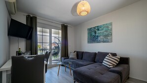 A modern living room with a dark grey sofa, a dining table with chairs, a flat-screen TV mounted on the wall, and a large window with curtains. - Apartments Domic (Medulin)