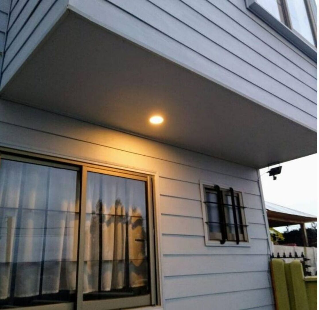 Ett hus med en inglasad veranda, ett stort fönster med gardiner och ett mindre fönster med en fönsterlucka.