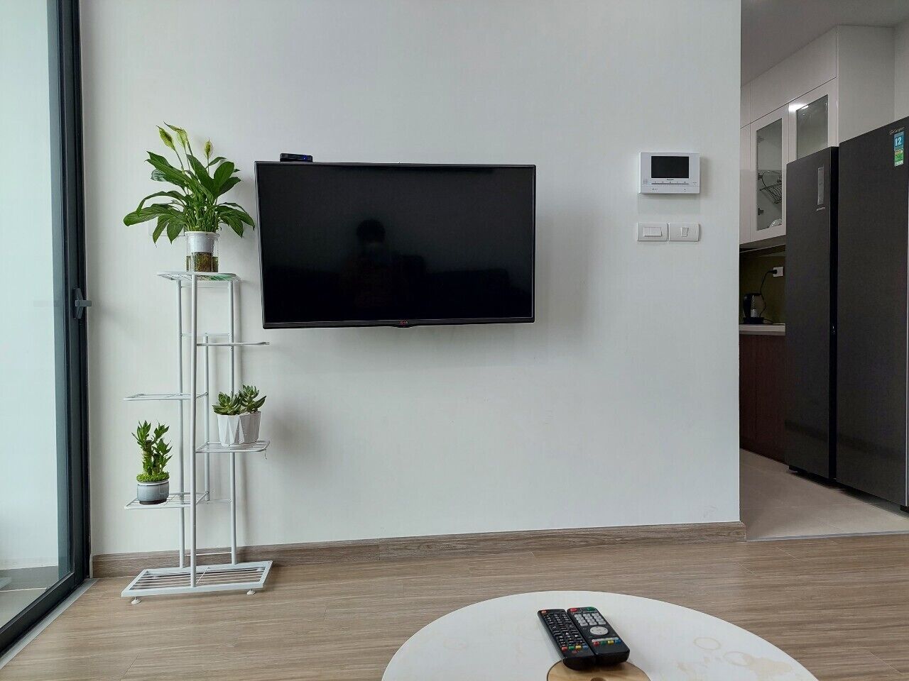 Een moderne woonkamer met een flatscreen televisie aan de muur, een witte plantenstandaard met meerdere potplanten, een afstandsbediening op een witte ronde tafel, en een keuken in de achtergrond.