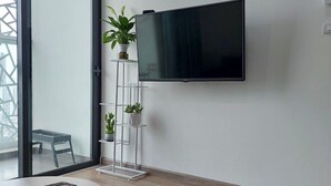 Una sala de estar moderna con un televisor de pantalla plana grande montado en la pared, una mesa de centro redonda con un control remoto, y un soporte para plantas con varias macetas.