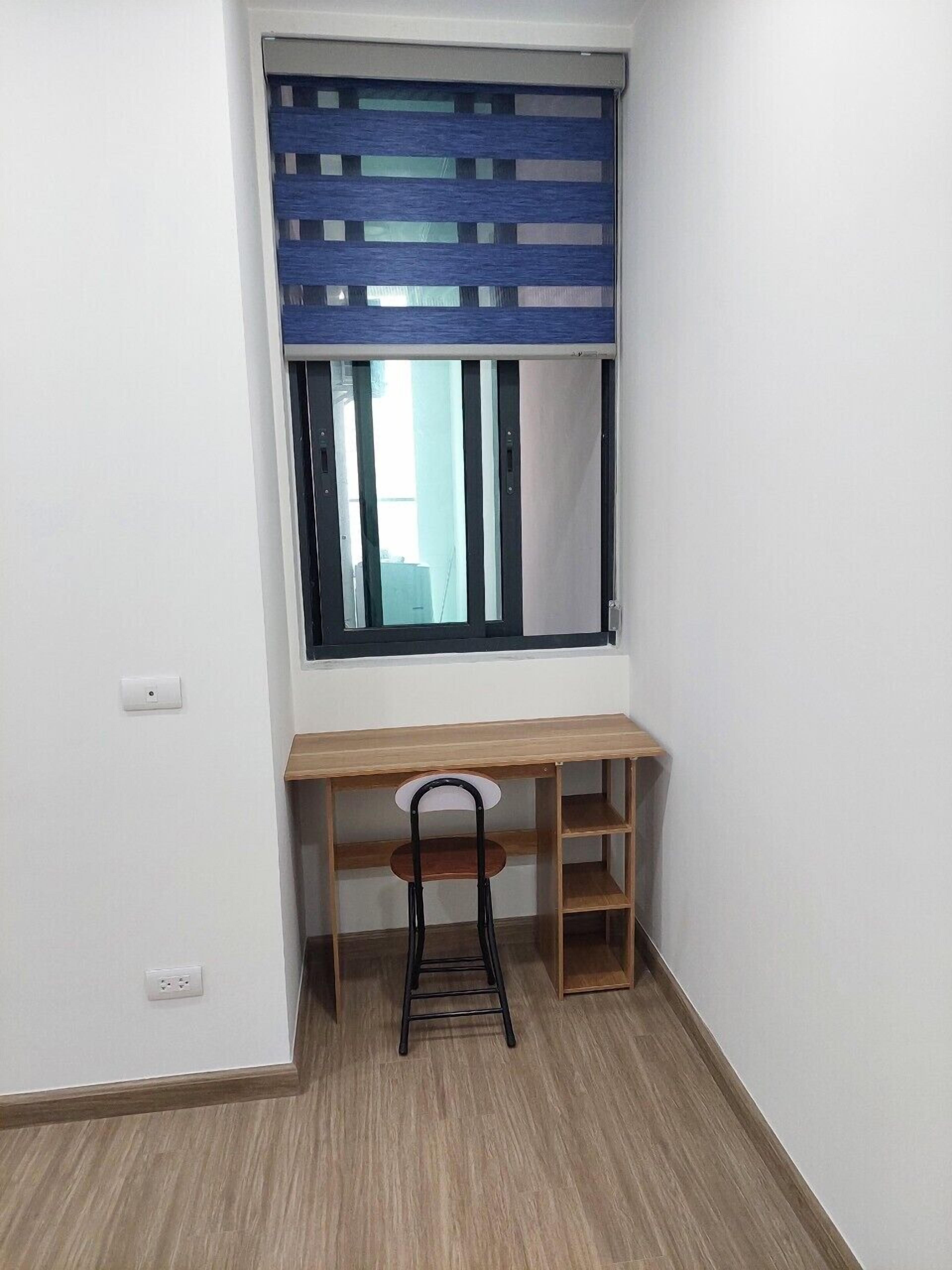 A corner room with a wooden desk, a chair, a window with blue blinds, and a light switch on the wall.
