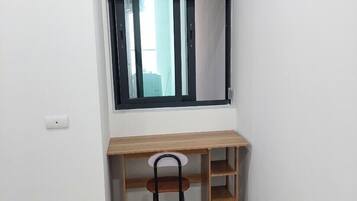 A corner room with a wooden desk, a chair, a window with blue blinds, and a light switch on the wall.
