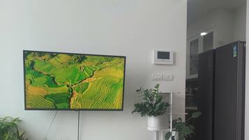 En platt-TV monterad på en vit vägg, en liten termostat och en krukväxt på ett vitt stativ.