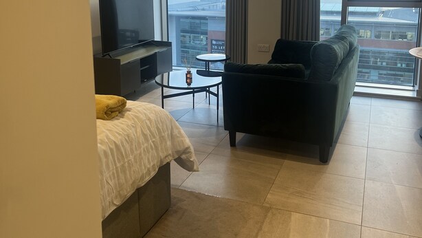 A modern hotel room with a bed, a sofa, a TV, a small table, and large windows offering a city view.
