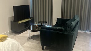A modern living room with a dark green sofa, a flat-screen TV on a black stand, a small round table with a candle, and grey curtains.