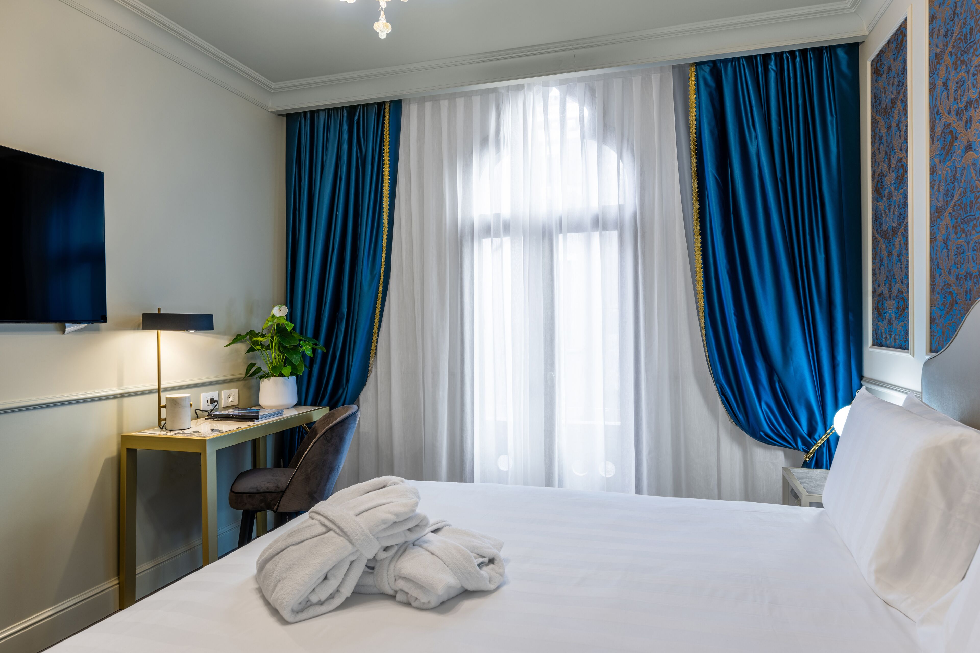 A hotel room with a bed, a desk, a chair, a TV, and a window with blue curtains.