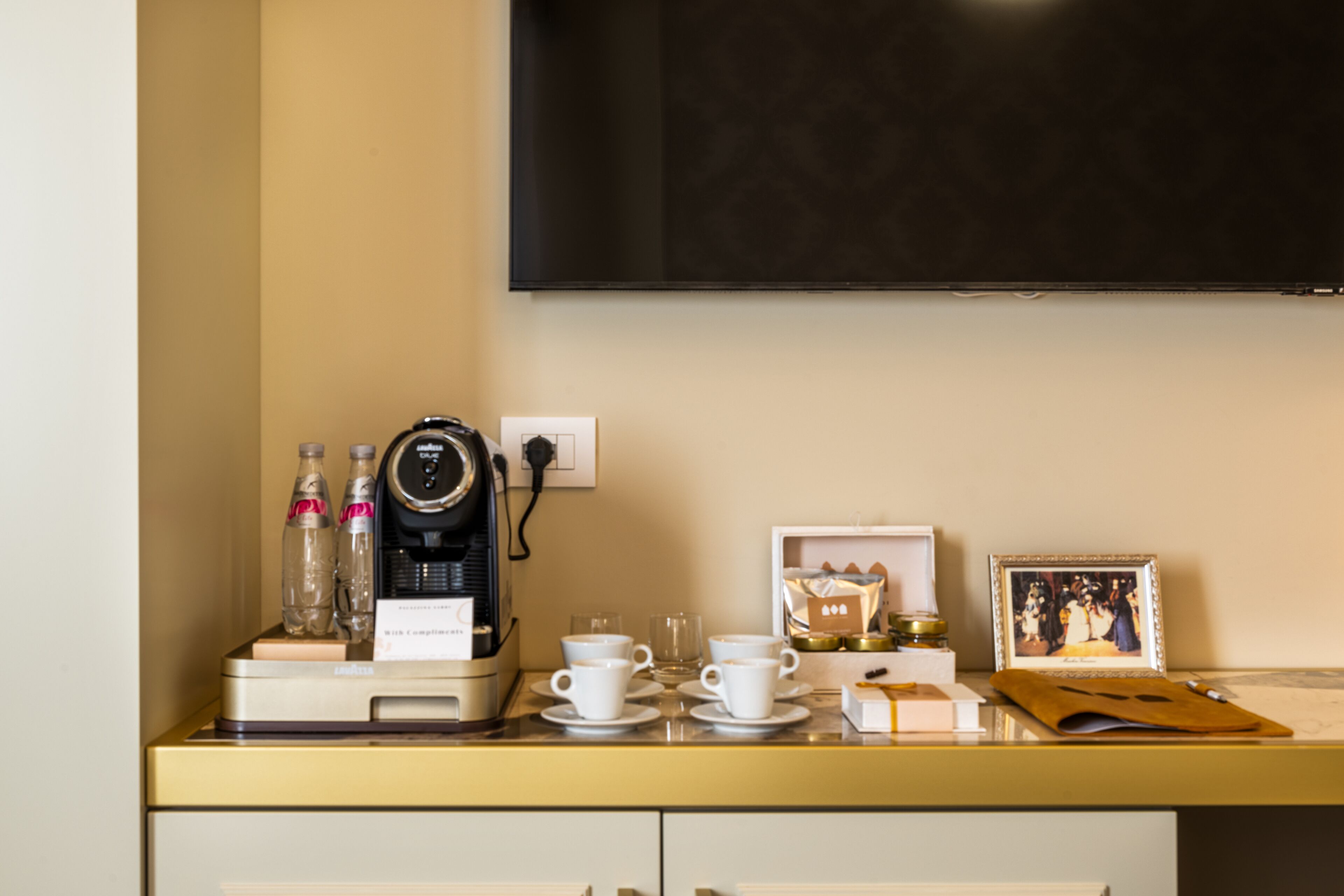 A hotel room with a minibar, a coffee machine, cups, a framed photo, and a television on the wall.
