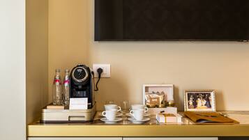 A hotel room with a minibar, a coffee machine, cups, a framed photo, and a television on the wall.