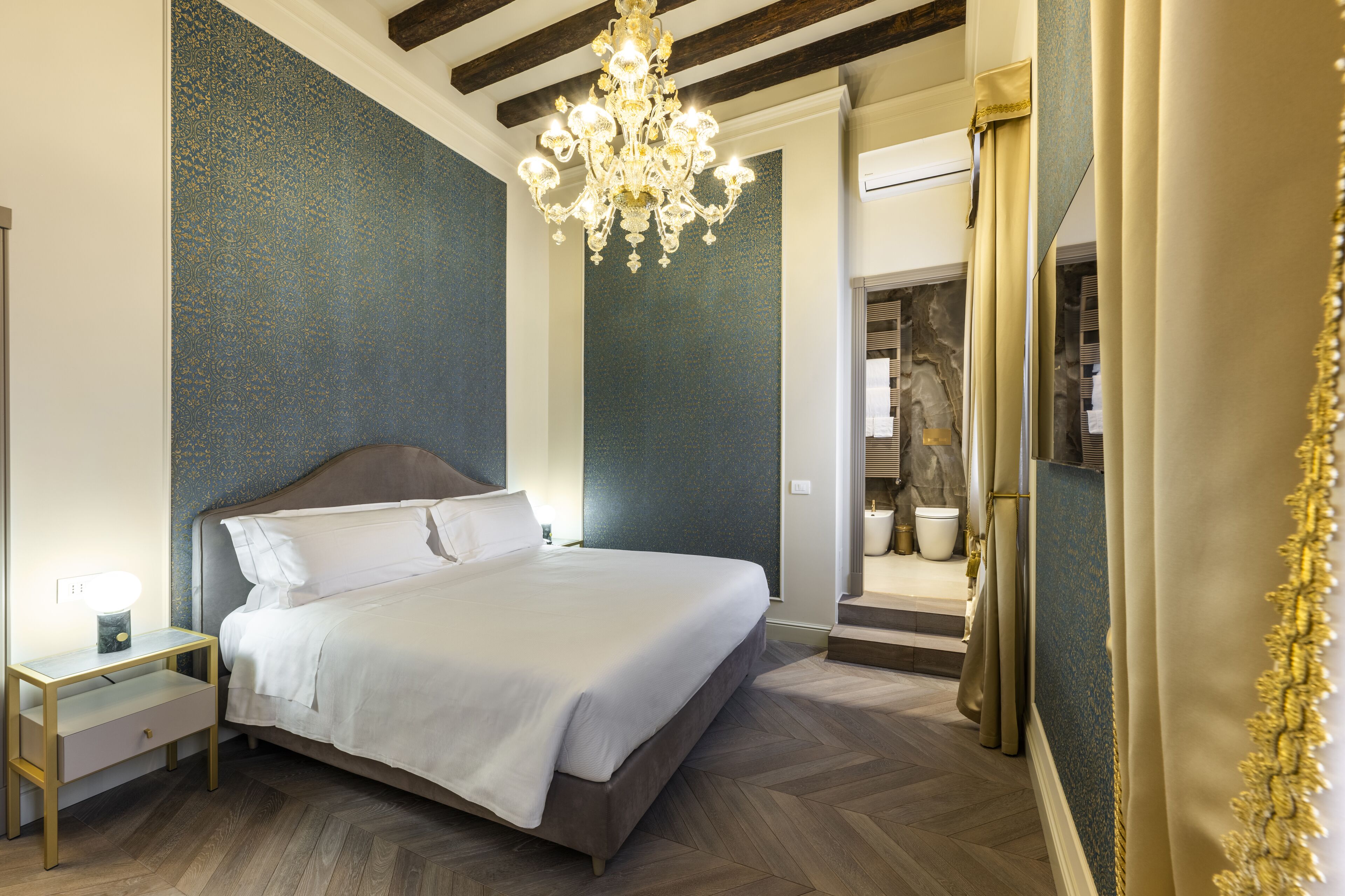 Une chambre à coucher avec un grand lit, une table de chevet, un lustre et une salle de bain visible à travers une porte ouverte.
